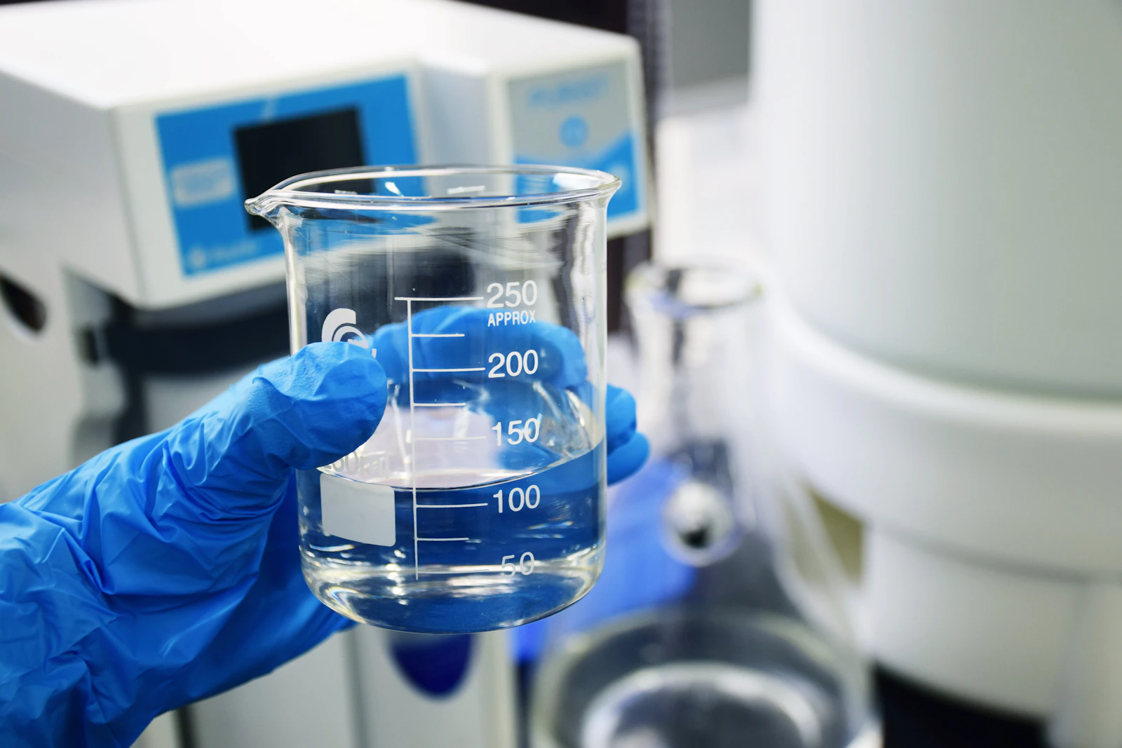 Mano con guante azul sosteniendo un vaso de laboratorio con líquido transparente.